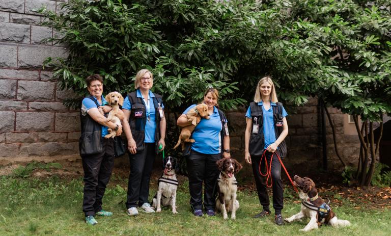 The canines for care team posing with their dogs in front of a brick wall and trees