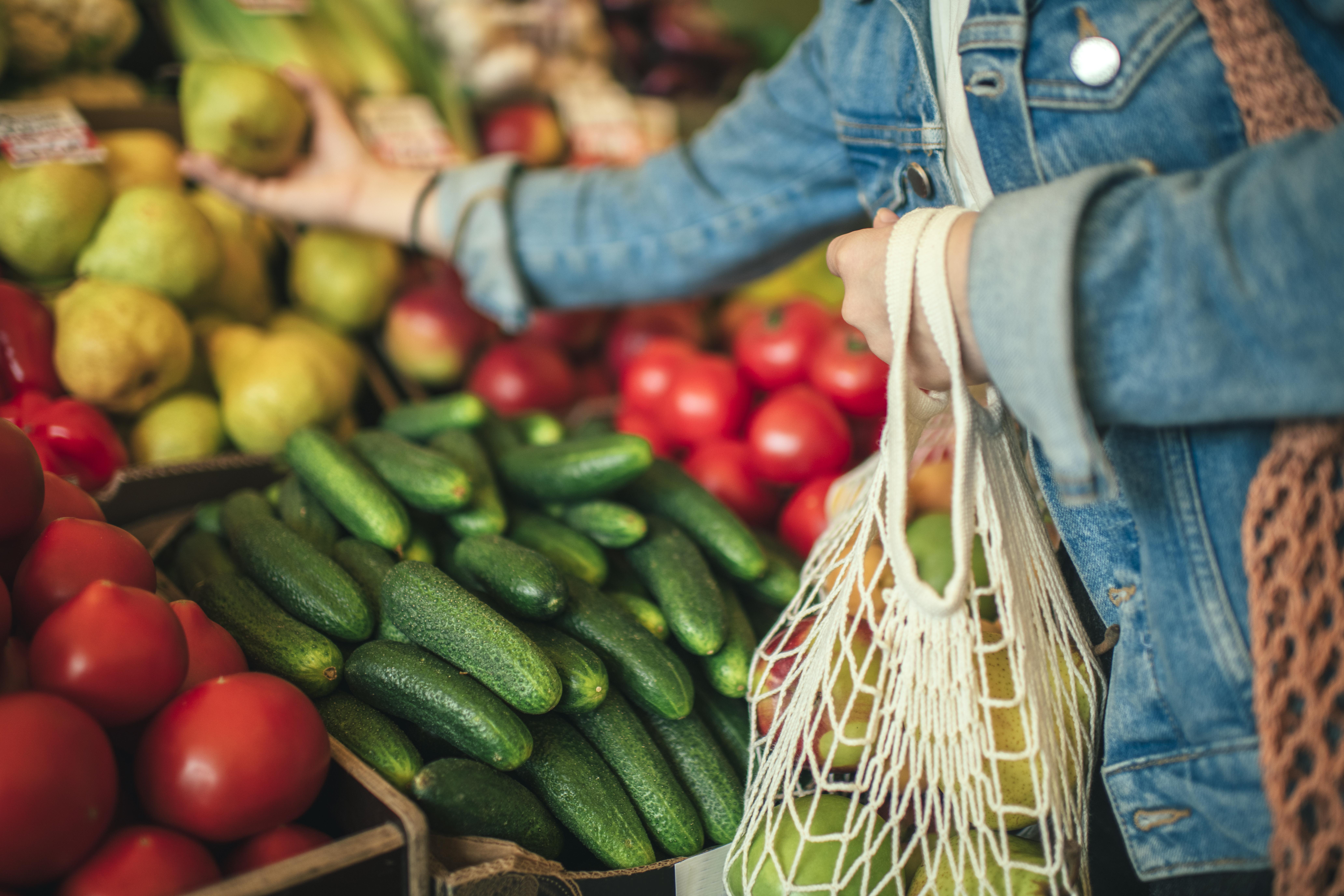 Person at a farmers market selecting fruits and vegetables using a reusable bag
