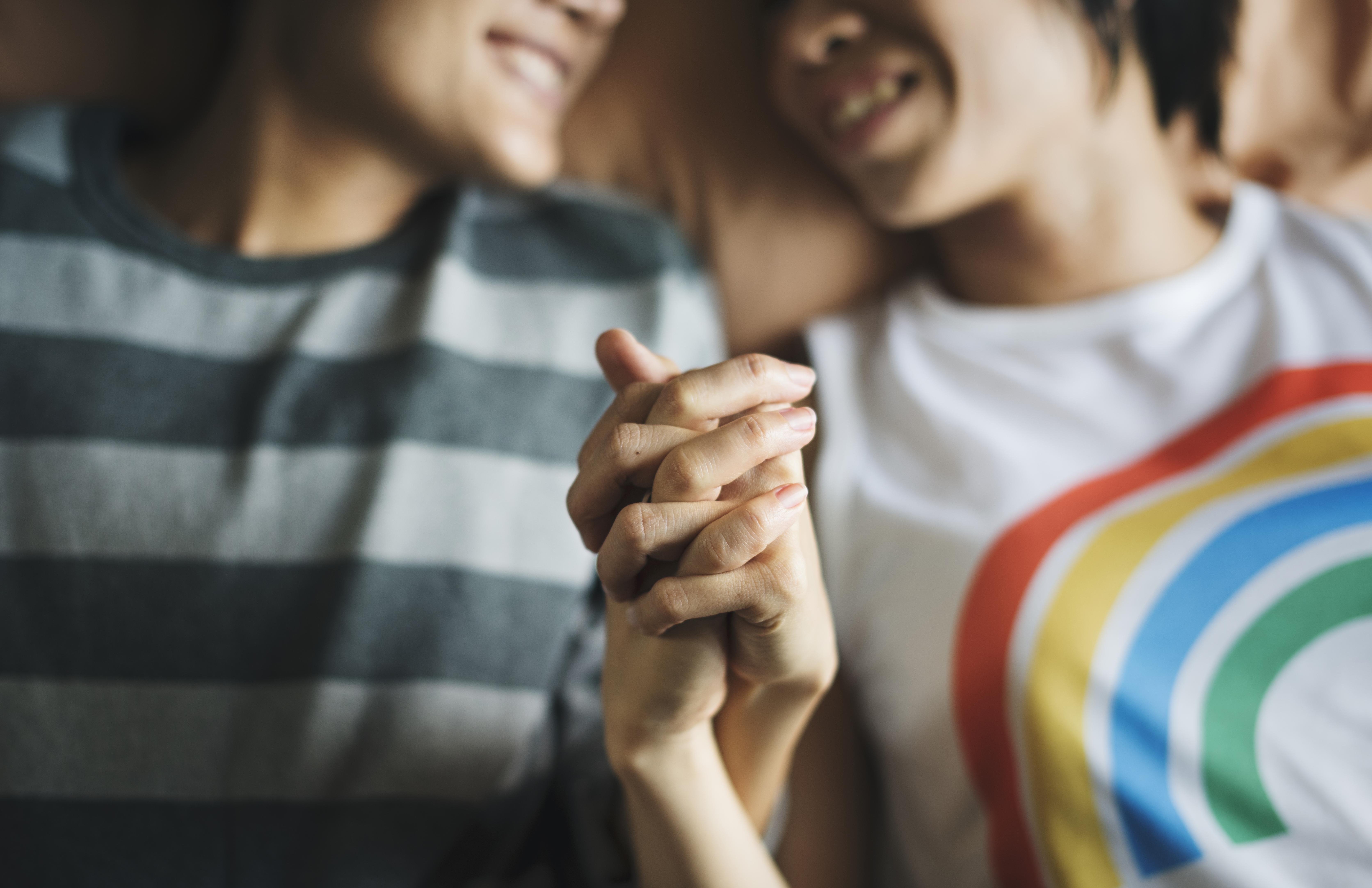 Closeup of two people laying on a bed holding hands, one of them is wearing a rainbow tshirt